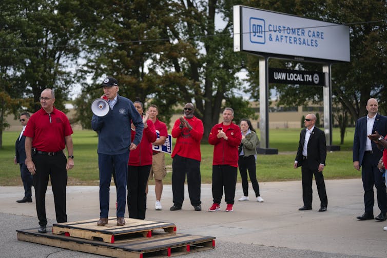 President Biden, in blue, speaking into a megaphone near several people dressed in red in front of signs saying GM and UAW.