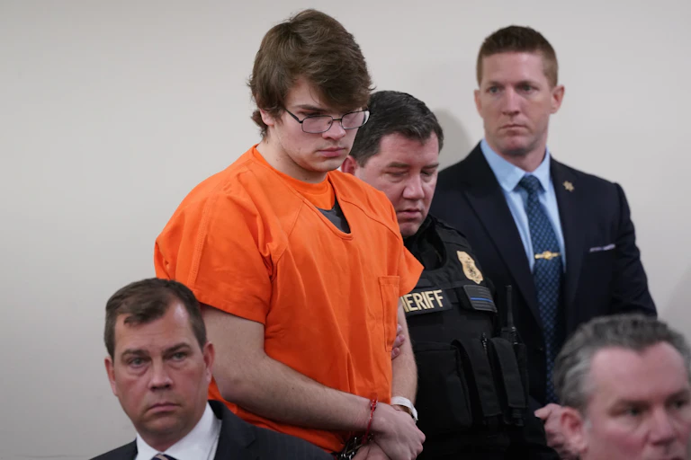 A young white man wearing an orange shirt is handcuffed and surrounded by law enforcement officers.