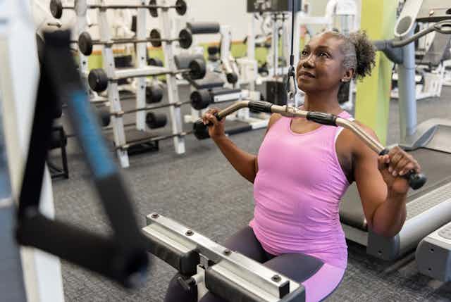 Middle-aged woman wearing athletic clothing pulls down on a weighted bar in a gym. 