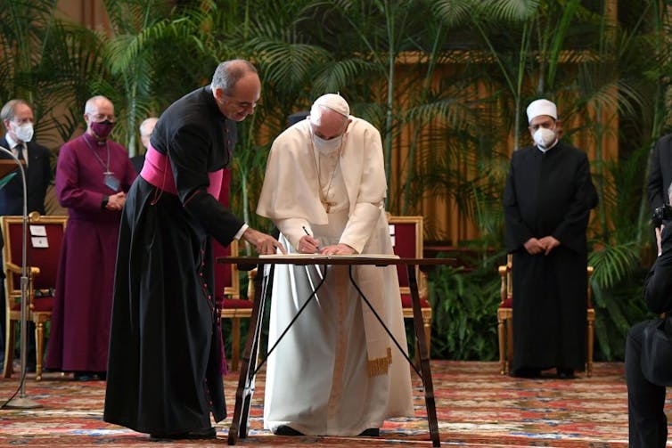 A man in white robes bends over a small table to sign something, as men in black and purple robes stand behind him.