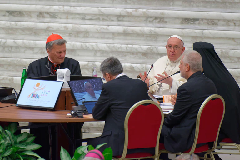Man in white robe speaking into a microphone while seated at a round table with four other men, all with a computer screen in front of them