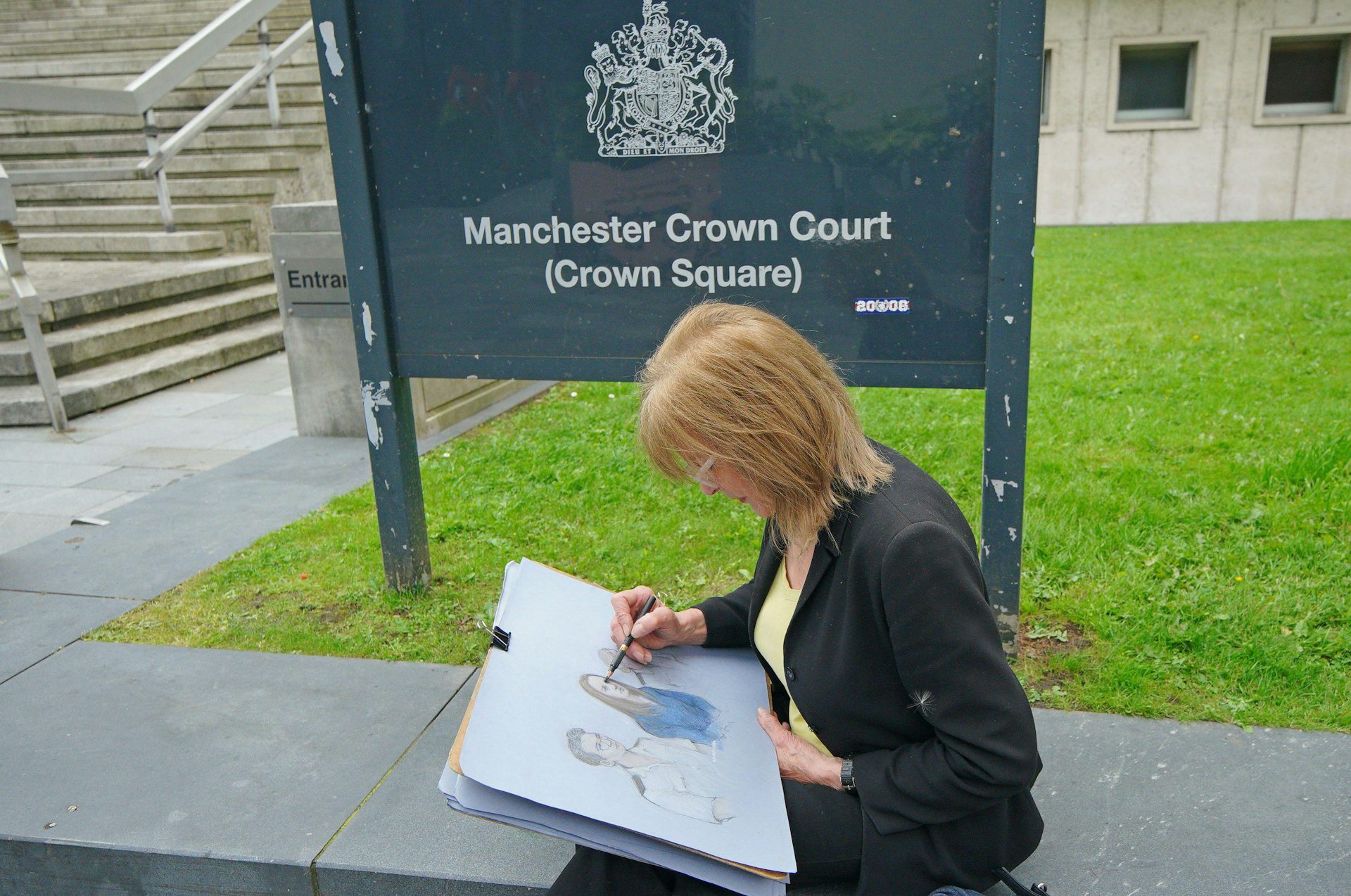 Woman with drawing pad sits on a wall in front of sign for Manchester Crown Court.
