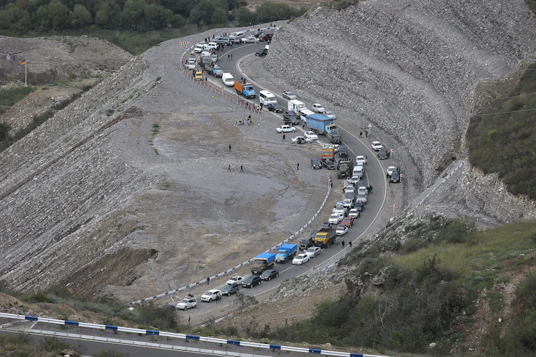 A convoy of cars along a winding highway amid gravelly hills.