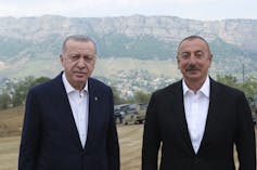Two men in dark jackets and white shirts pose for a photo with mountains behind them.