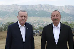 Two men in dark jackets and white shirts pose for a photo with mountains behind them.