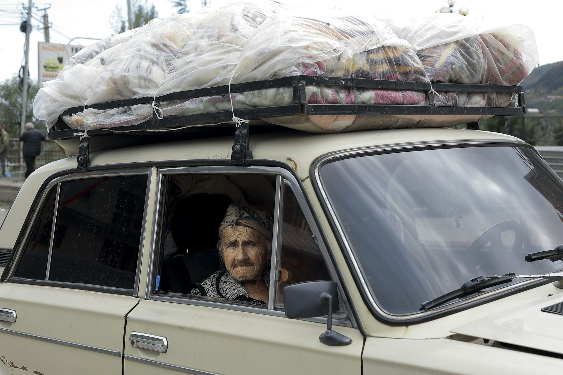 An elderly woman sits in a car laden with belongings on its roof.