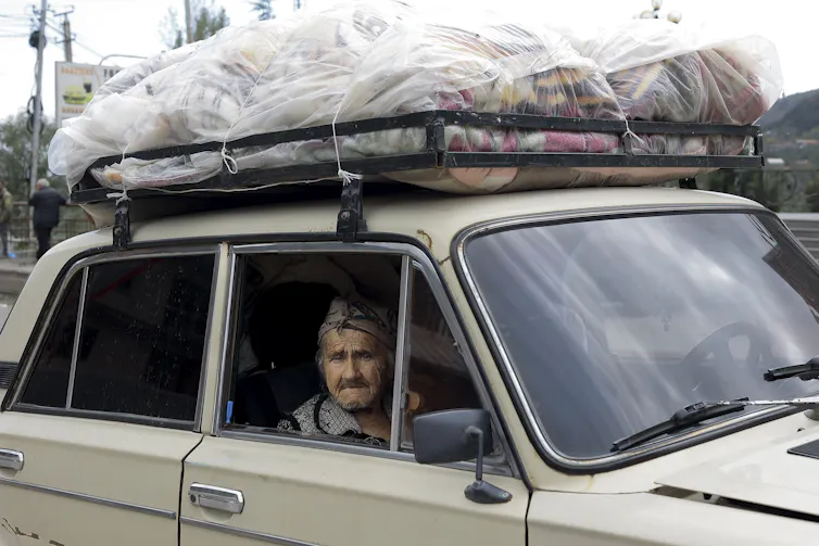 An elderly woman sits in a car laden with belongings on its roof.