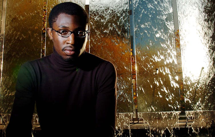Young Black man wearing a black turtleneck and eyeglasses posing in front of a sculpture with waterfalls.