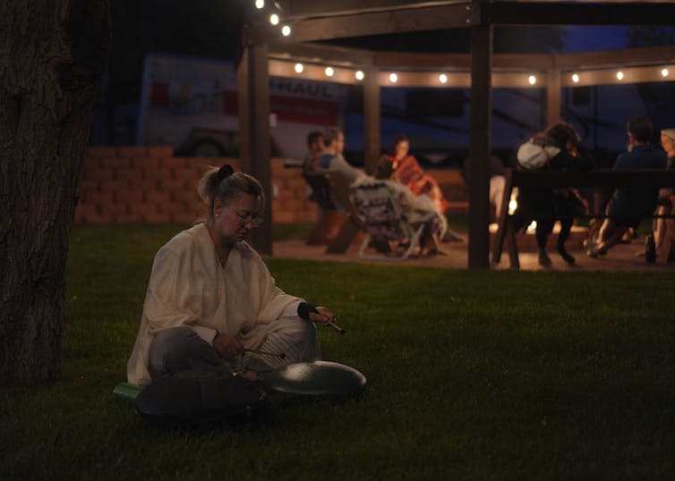 A woman seated near a tree, on grass, plays a sound bowl.