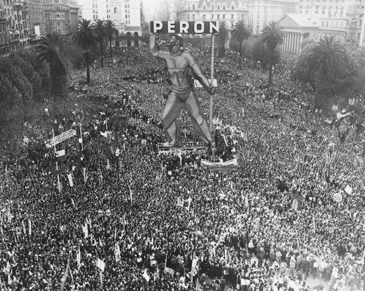 A crowd of people stand around a large figure with 'PERON' written at the top.