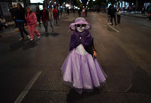 Little girl wearing a pink dress and a purple shawl, with her face painted like a skull.