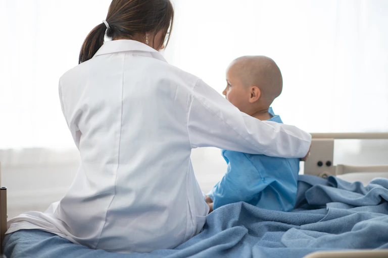 Doctor with arm around the shoulders of a young cancer patient, both sitting on a hospital bed