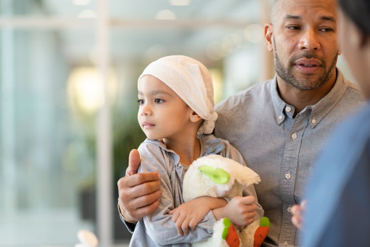 Father carrying child with cancer wearing a bandana and holding a stuffed animal, talking to a health care provider