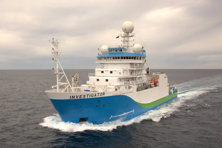 Photo of a blue-and-white ship, the RV Investigator, sailing through the sea beneath grey skies.