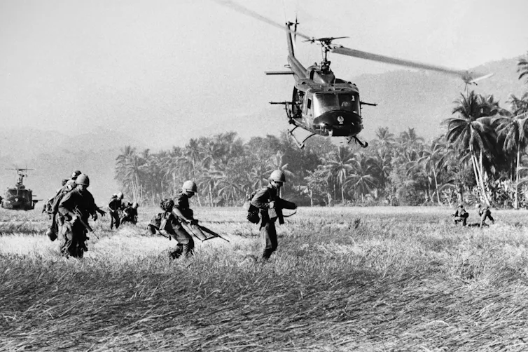 Black and white image of US soldiers in south Vietnam with helicopters circling above.
