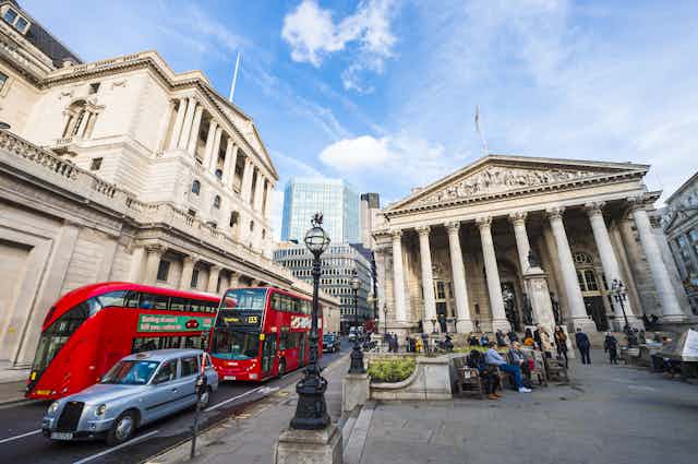 Taxis and buses pass the Bank of England building while people sit outside the Royal Exchange building in London, blue sky. 