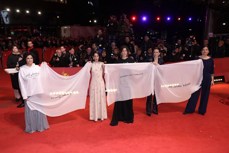 Mujeres elegantemente vestidas en una alfombra roja sostienen una pancarta, con una fila de fotógrafos detrás de ellas.