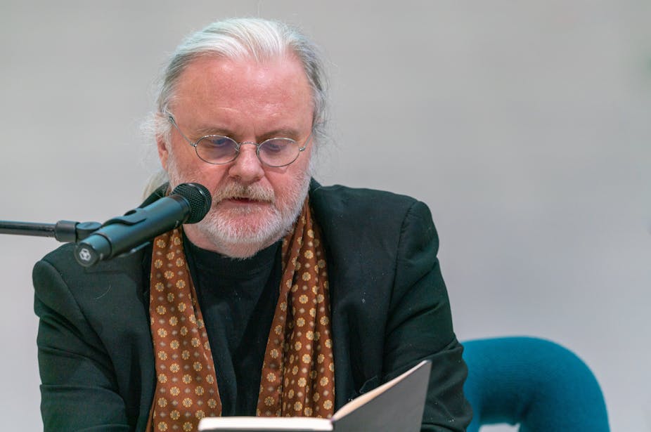 A grey haired bespectacled and bearded man with a scarf round his neck, reading from his book into a microphone.