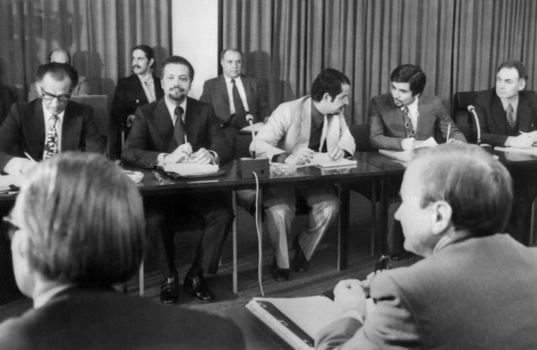 Men in suits sit at two rows of tables across from one another. Ahmed Zaki Yamani is looking into the camera.