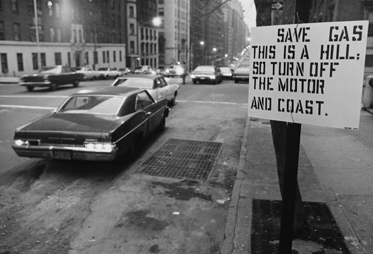 A large car drives on a street in New York. A sign reads 'Save gas. This is a hill, so turn off the motor and coast.'