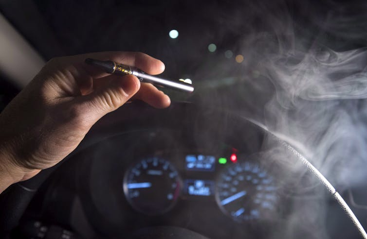 Close-up of a hand holding a smoking cannabis oil vaporizer in front of a steering wheel and dashboard