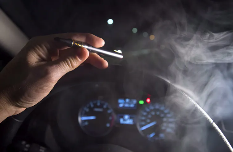 Close-up of a hand holding a smoking cannabis oil vaporizer in front of a steering wheel and dashboard