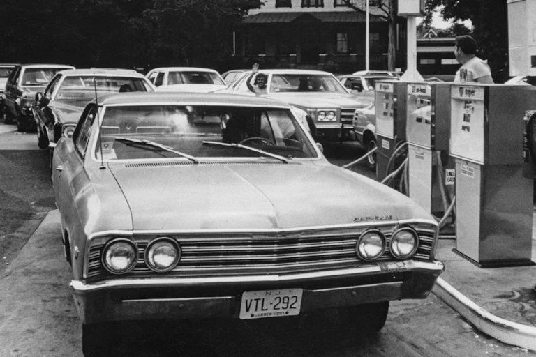 Large cars line up for gas gasoline in a long line. A man waves from a car window.