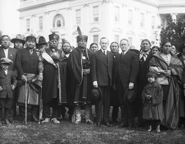 Men and women in a mix of suits and traditional dress stand in front of the White House
