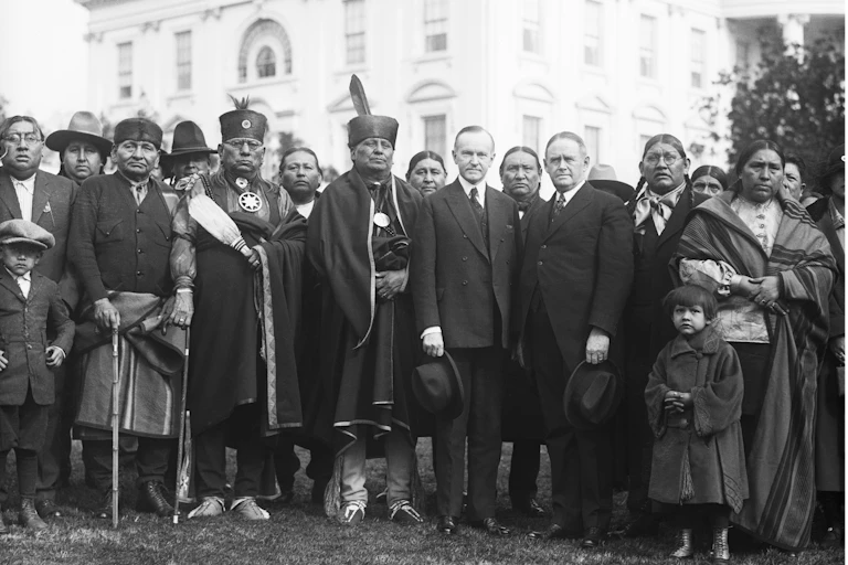 Men and women in a mix of suits and traditional dress stand in front of the White House