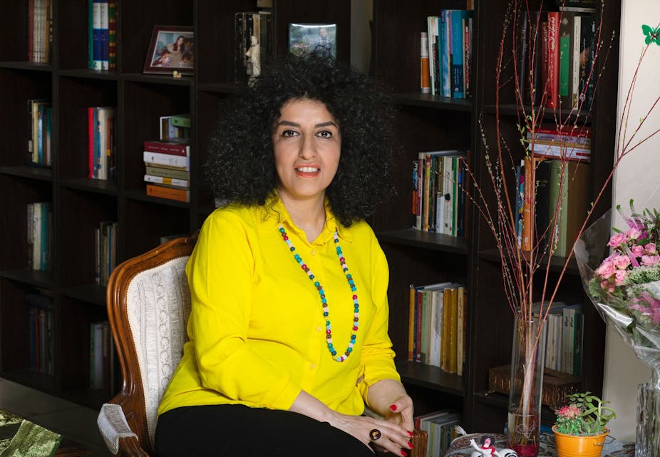 A woman wearing a yellow shirt and long necklace sitting on a chair, set against a large bookcase.