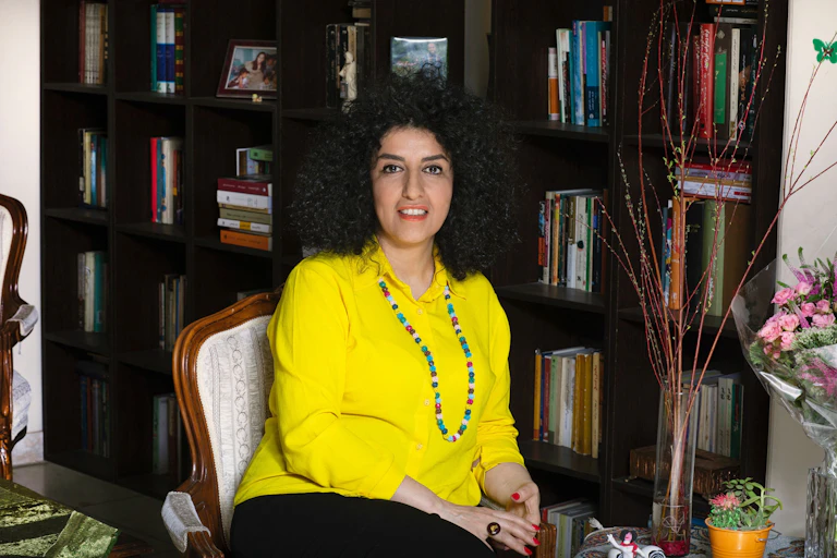 A woman wearing a yellow shirt and long necklace sitting on a chair, set against a large bookcase.