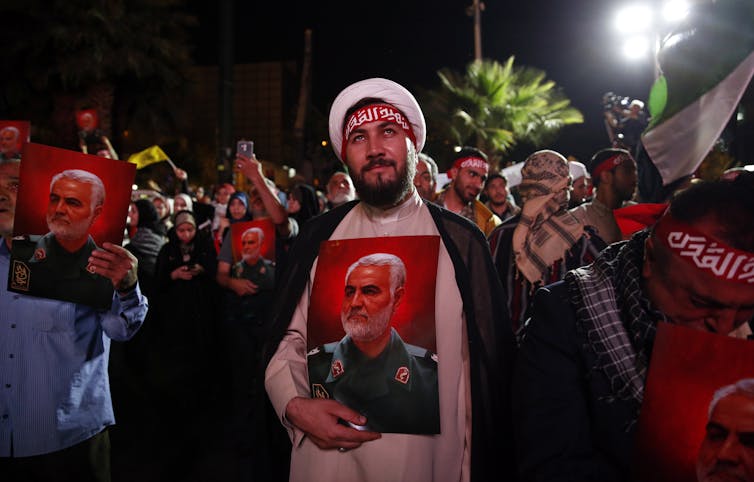 Man in Iranian dress carries portrait of a solider, in front of a crowd of people in Islamic dress.