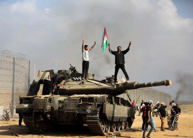 Dois homens, um empunhando a bandeira palestina, estão de pé sobre um tanque de guerra israelense