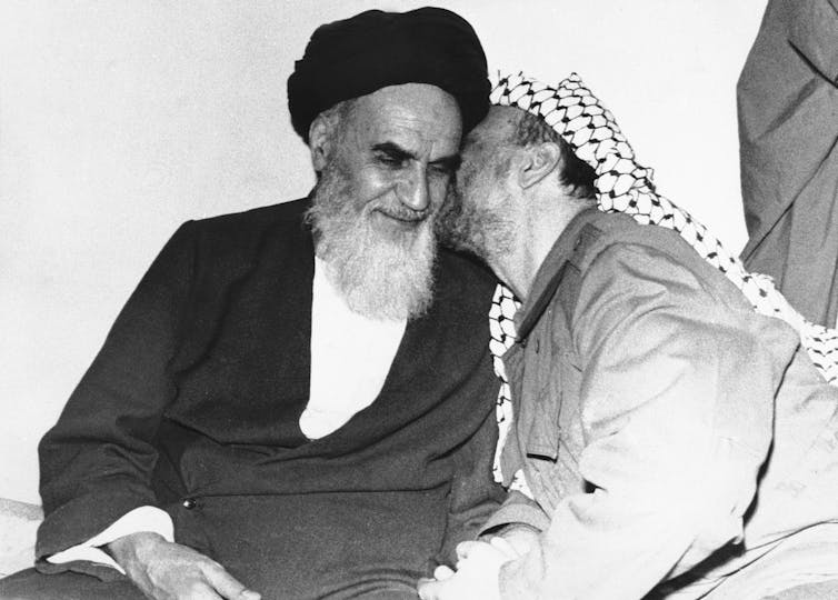 A man in a black robe and head covering is kissed on the cheek by a man wearing a black and white head covering.