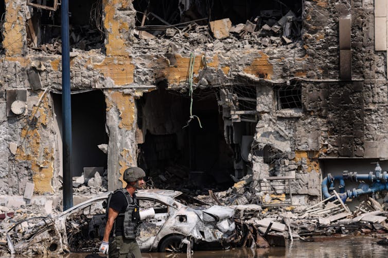 A man in a camouflage helmet walks past the broken facade of a building