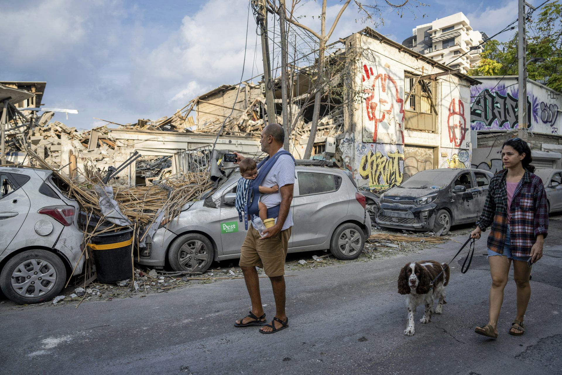 A man holding a baby and a woman with a dog look at bombed out buildings and cars