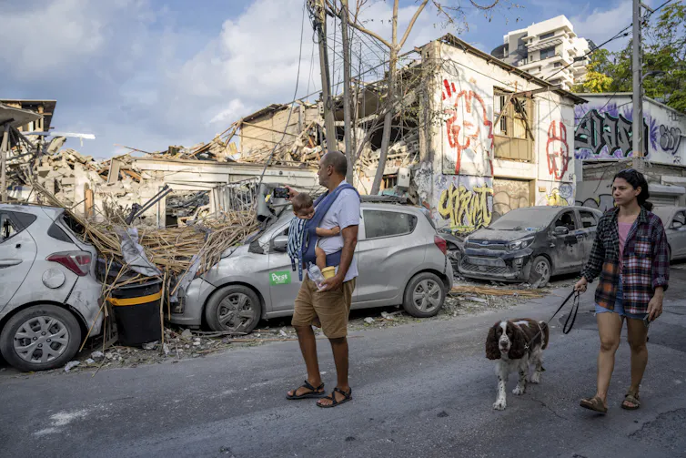A man holding a baby and a woman with a dog look at bombed out buildings and cars