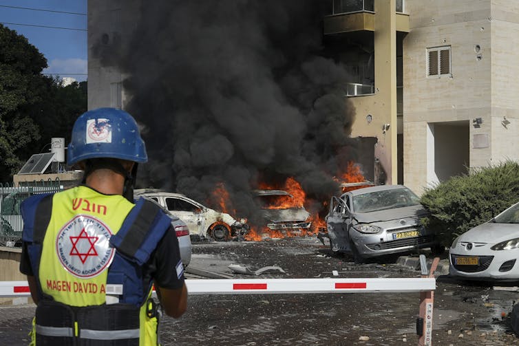 A man in a helmet and protective vest looks on as cars burn in the background.
