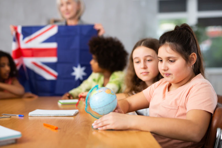 A child is looking at a globe with a teacher in the background holding an Australian flag.