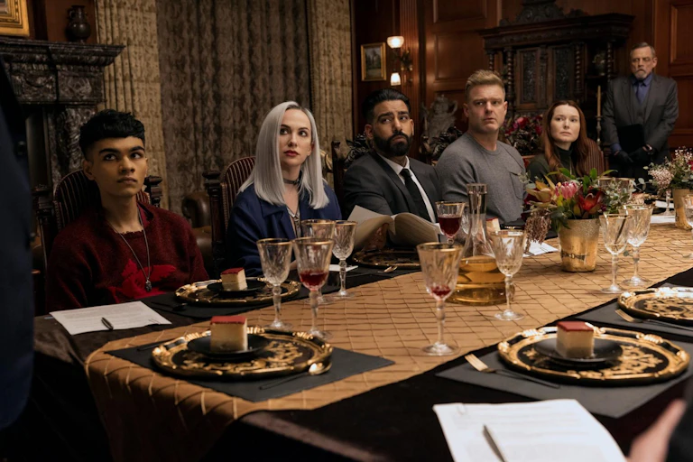 Cinco personas se sientan en una mesa de comedor profusamente decorada y miran a alguien que se encuentra fuera de campo mientras una sexta persona espera de pie, al fondo.