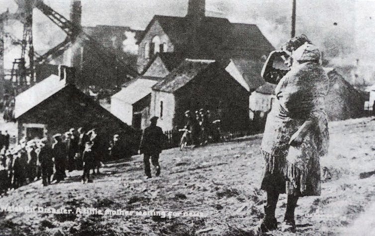 A black and white image depicting a woman with a baby on the side of a hill. A coal mine is seen in the distance.