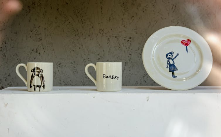 Two mugs and a plate with poor representations of Banksy works on them.