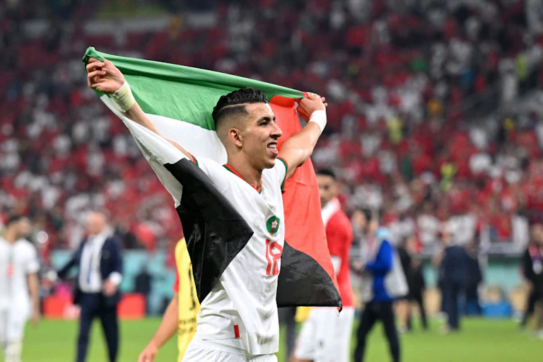 A man with shaven hair holds a flag aloft as he smiles, running across the field of a sports stadium.