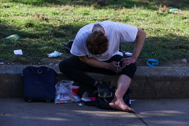A white man is slumped over on a curb, holding a needle in his hand. Garbage surrounds him.