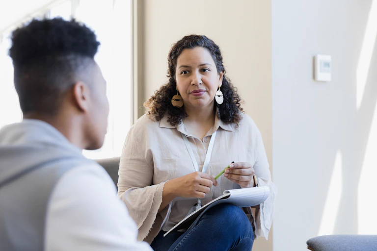 A teen boy talks with a counselor who has a notepad and pen. They are both seated.