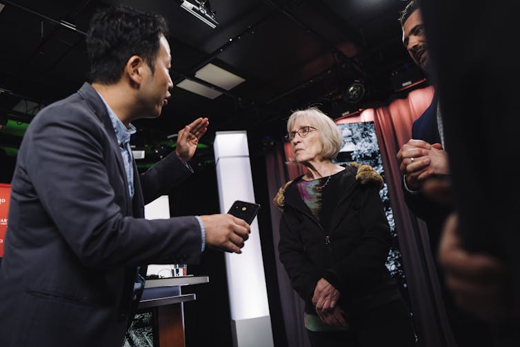 A young reporter in a suit is shown speaking to economist Claudia Goldin, who stands with her hands clasped.