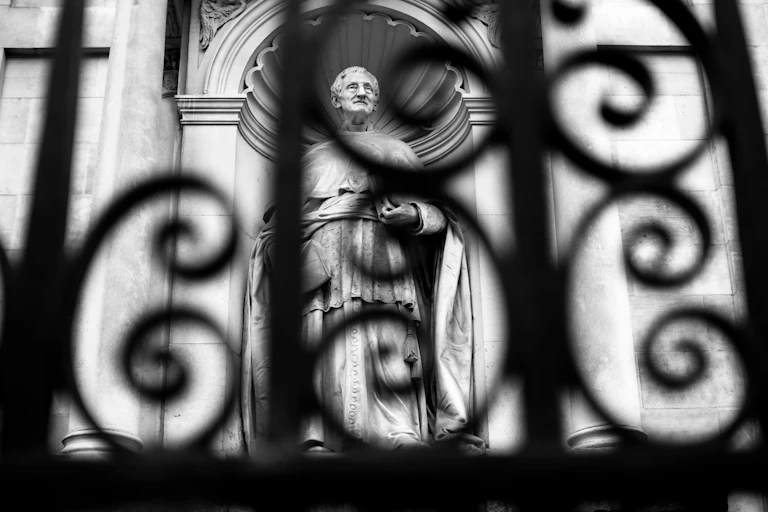 A black and white photo of a statue shot from behind an elaborate iron gate.