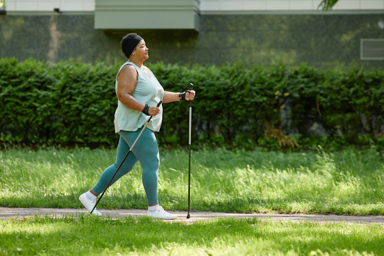 Une femme ronde de profil, en tenue de sport, marche avec un bâton main. Elle pratique la marche nordique.