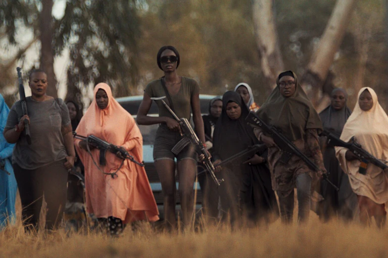 Women carrying guns walk in a row in front of a car, in the middle a tall young woman in sunglasses.