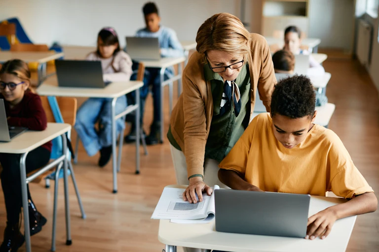 Profesor ayudando a un alumno a utilizar un ordenador en clase.